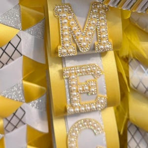May include: A decorative homecoming mum accessory featuring yellow and white satin ribbons, silver glitter accents, and a row of faux pearl floral embellishments. The letters M, E, C, O are displayed vertically, each crafted with white faux pearls.