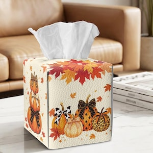 May include: A square tissue box cover with a textured cream background featuring a pattern of orange pumpkins, black and white bows, and red and orange autumn leaves. A stack of white tissues protrudes from the top opening of the box.