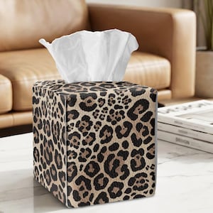May include: A square tissue box cover featuring a tan and black leopard print pattern. The exterior has a textured surface, and a single white tissue is pulled through the top opening, extending upward from the center of the box.