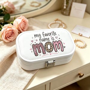 May include: A white rectangular jewelry box with a silver metal latch. The lid features the black text "my favorite name is" above the word "mom" written in large, pink glittery letters with a leopard print pattern and small pink hearts.