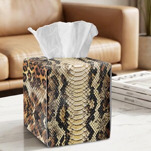 May include: A square tissue box cover featuring a detailed snakeskin pattern in shades of brown, tan, and black, with a leopard print design on the side panels. A stack of white tissues protrudes from the top opening of the box cover.