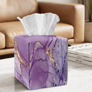 May include: A square tissue box cover featuring a purple marble pattern with gold veining accents. The exterior has a textured finish, and a white tissue is pulled through the top opening. The design displays deep violet and light lavender hues.