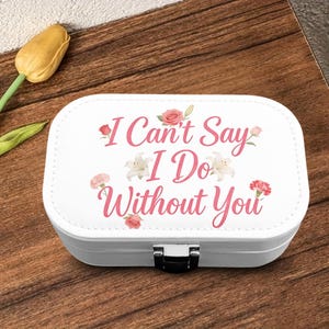 May include: A white rectangular jewelry box with rounded corners and a silver metal latch. The lid features the pink script text I Can't Say I Do Without You, surrounded by illustrations of pink roses, carnations, and white lily flowers.