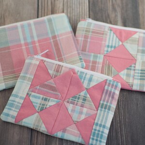 May include: Three rectangular zipped pouches made of fabric. One pouch features a simple pink, blue, and white plaid pattern. The other two pouches display a patchwork design combining solid pink fabric triangles with plaid fabric squares.