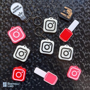 Peut inclure: Divers porte-clés et breloques, dont des logos Instagram rouges et blancs, une ampoule avec le texte "LUMINE ELECTRICAL", et un charme de vernis à ongles. Ils sont disposés sur une surface sombre et texturée.