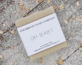 Może przedstawiać: Naturalne mydło w kostce z białą etykietą, na której widnieje napis "Steamboat Soap Company" i "Oh Baby" czarnym kolorem. Etykieta zawiera również napis "All Natural Soap" i "Handmade in Steamboat Springs, CO".