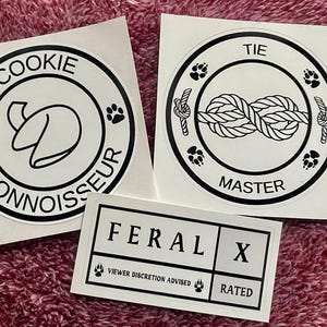 May include: Three white stickers with black text and graphics. The first sticker says "Cookie Connoisseur" with a fortune cookie graphic. The second sticker says "Tie Master" with a knot graphic. The third sticker says "Feral X" with the text "Viewer Discretion Advised" and "Rated" below.