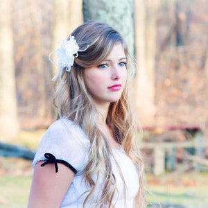 K&ouml;nnte beinhalten: Eine junge Frau mit langen blonden Haaren tr&auml;gt ein wei&szlig;es Kleid mit einem schwarzen Band am &Auml;rmel. Sie hat eine wei&szlig;e Blume im Haar und schaut zur Seite.