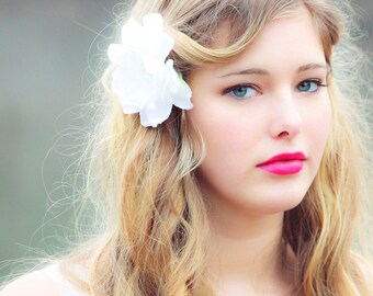 Puede incluir: Una joven con el pelo largo y rubio peinado en ondas. Lleva un vestido blanco y tiene una flor blanca en el pelo. Mira a la cámara con una sonrisa suave.