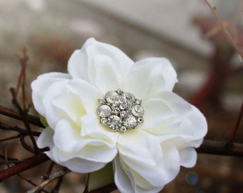 Puede incluir: Una flor de tela blanca con un grupo de pedrería transparente en el centro. La flor tiene múltiples pétalos y está rodeada de ramitas marrones, creando un aspecto natural y elegante.