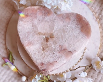 May include: A heart-shaped pink and white agate geode with a crystal cluster inside. The geode is sitting on a white marble pedestal with gold accents.