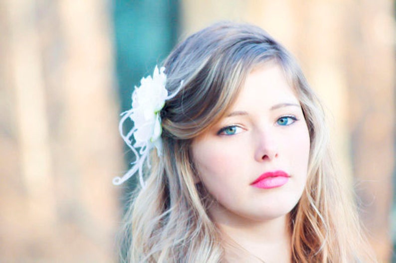 K&ouml;nnte beinhalten: Eine junge Frau mit langen blonden Haaren, die mit einer wei&szlig;en Blume hinter dem Ohr gestylt sind. Sie tr&auml;gt leichtes Make-up und hat einen sanften, nat&uuml;rlichen Look.