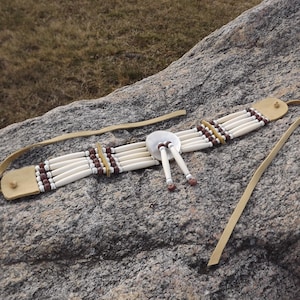 May include: A handcrafted choker necklace featuring off-white bone beads, brown and yellow accent beads, and a central silver-colored disc. The necklace has leather straps and is displayed on a rock surface. This piece is a unique accessory.