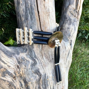 May include: A handcrafted necklace featuring a combination of natural materials. The necklace has a central, circular, brass-colored element with a cluster of silver-toned beads. Black, cylindrical beads are connected to the central piece, and the necklace is set against a natural wood backdrop.