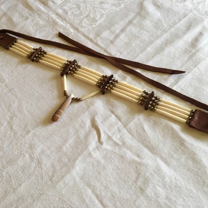 May include: A handcrafted choker necklace featuring off-white cylindrical beads, accented with bronze-colored beads and dark brown leather details. The necklace has a pendant with a wooden element and a brown leather tie.