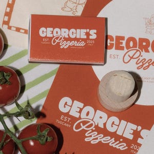 May include: An overhead shot features a matchbox and a coaster with the text "GEORGIE'S Pizzeria" in white on an orange background. Fresh red tomatoes and a wooden object are also visible. The matchbox reads "EST. TUSCANY 2025 ITALY."