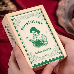 以下が含まれることがあります： 「BOOKLOVERS」の文字と本を読む女性のイラストが描かれた、緑と白のラベルが付いた木製のマッチ箱。赤いベルベットの背景に、手で箱が持たれています。「Book Reading Club」の文字も表示されています。