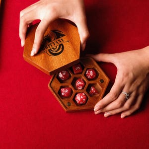 Puede incluir: Una caja hexagonal de madera con una tapa que presenta un diseño de dragón grabado con láser y el nombre Brittany. La caja abierta contiene siete dados poliédricos rojos con números blancos, cada uno en un compartimento hexagonal.