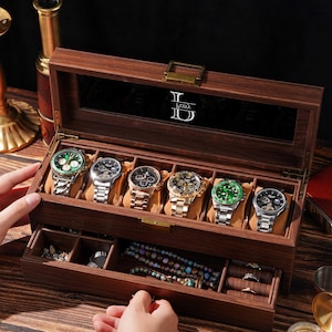 May include: A dark wood watch box with a glass lid featuring the white text "Perry". The interior has six watch slots with tan cushions and a bottom drawer containing compartments for jewelry, including rings and beaded bracelets.
