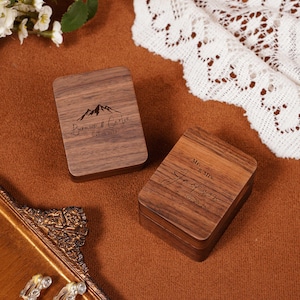 May include: Two wooden ring boxes with engraved names and dates. One box features a mountain graphic and the names "Bernice & Carlin" with "May 02, 2025." The other box says "Mr. & Mrs. Jackson" with "05.20.26."