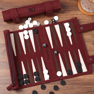 May include: A burgundy suede backgammon set featuring a roll-up board with dark brown and cream triangular points. The board displays the name Camila, while the matching storage case is personalized with the initials SG. Includes black and white checkers and dice.