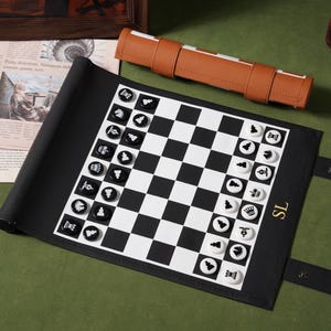 Op de afbeelding: Een oprolbaar schaakbord van zwart leer met een wit en zwart raster en ronde speelstukken. Het bord toont de letters SL in goud aan de rechterkant. Een aparte draagtas van bruin leer is boven het bord geplaatst.