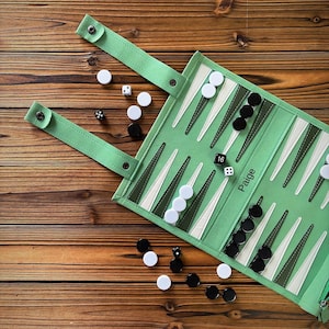 Puede incluir: Un juego de backgammon enrollable de tela verde claro con puntos triangulares en verde oscuro y blanco. El tablero tiene el nombre Paige impreso en negro. Incluye fichas circulares blancas y negras, dados y dos correas con cierre de botón a presión.