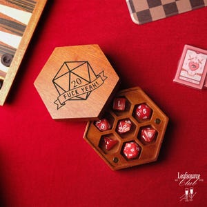 Könnte beinhalten: Sechseckige Würfelbox aus Holz mit dem Text "20 FUCK YEAH!" und einer D20-Grafik. Die Box ist geöffnet und zeigt rote Würfel. Die Würfel befinden sich in einem holzfarbenen, wabenförmigen Halter. Der Hintergrund ist rot.
