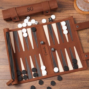 May include: A brown backgammon set with white, black, and gray accents. The set includes a zippered case, dice, and game pieces. The case is embossed with the initials "SG". The board is open, with pieces in play. The name "Camila" is written on the board.