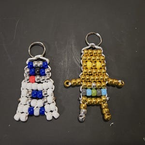 May include: Two beaded keychains, one white with blue and red beads, the other gold with yellow, blue, green, and gray beads.