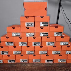 May include: A pyramid of orange shoe boxes, each with a label that reads "SARAH'S 1ST BIRTHDAY JULY 1, 2023 THANKS FOR BELIEVIN IT". The boxes are stacked on a dark wooden surface.