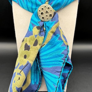 May include: A blue and teal floral patterned scarf with a decorative metal clasp. The scarf is draped over a mannequin head, showcasing its design and texture. The scarf's edges are trimmed with a teal border.