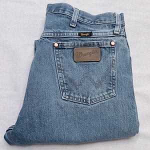 May include: Light blue denim Wrangler jeans. The jeans have a classic five-pocket design, a brown leather patch with the Wrangler logo, and copper rivets. The jeans are folded neatly.