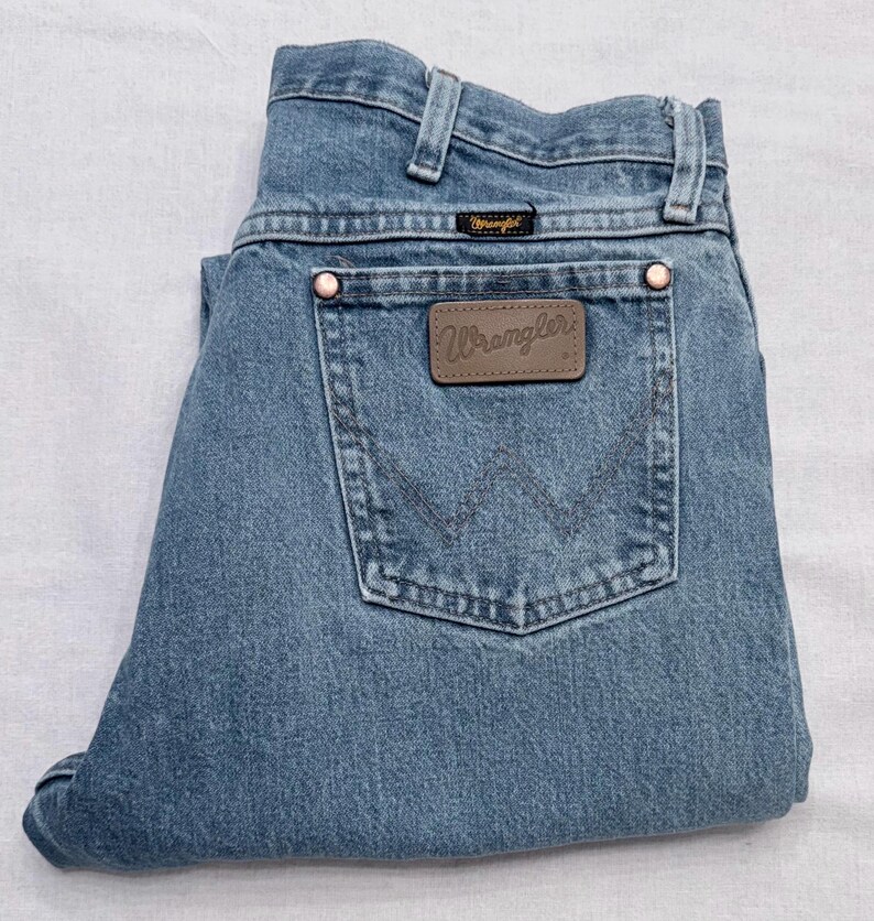 May include: Light blue denim Wrangler jeans. The jeans have a classic five-pocket design, a brown leather patch with the Wrangler logo, and copper rivets. The jeans are folded neatly.
