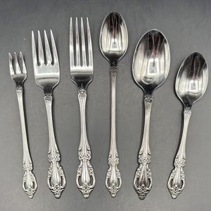 May include: Six pieces of silver silverware, including forks and spoons, are arranged on a dark surface. The silverware has ornate floral detailing on the handles. The set includes a small fork, two larger forks, and three spoons.