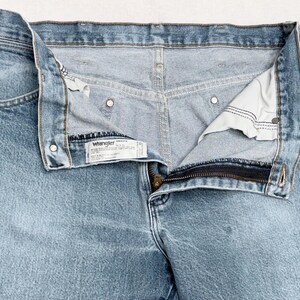May include: Light blue Wrangler jeans with a partially unzipped zipper. The jeans have a classic five-pocket design, metal rivets, and a Wrangler label. The denim fabric has a faded appearance, suggesting wear and washing.