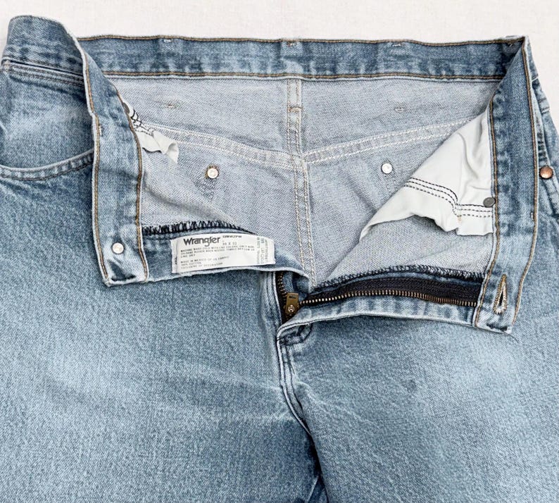 May include: Light blue Wrangler jeans with a partially unzipped zipper. The jeans have a classic five-pocket design, metal rivets, and a Wrangler label. The denim fabric has a faded appearance, suggesting wear and washing.