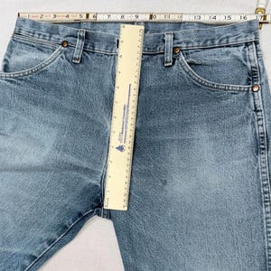 May include: Light blue denim jeans with classic five-pocket styling. The jeans are measured with a ruler, showing a waist measurement of approximately 16 inches across. Features include copper rivets and a button closure.