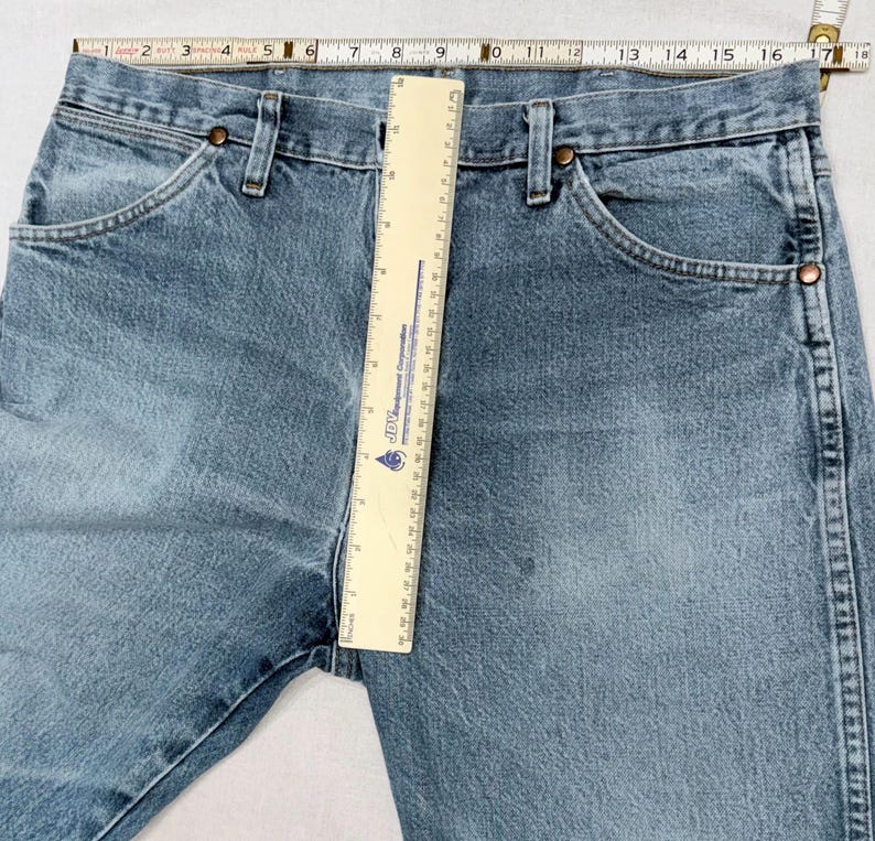 May include: Light blue denim jeans with classic five-pocket styling. The jeans are measured with a ruler, showing a waist measurement of approximately 16 inches across. Features include copper rivets and a button closure.