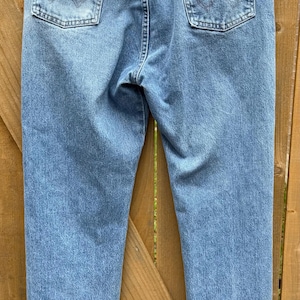 May include: Light blue denim jeans with classic five-pocket styling. The jeans feature a Wrangler brand patch on the back pocket and a metal button closure. The jeans are hanging on a hanger against a wooden background.
