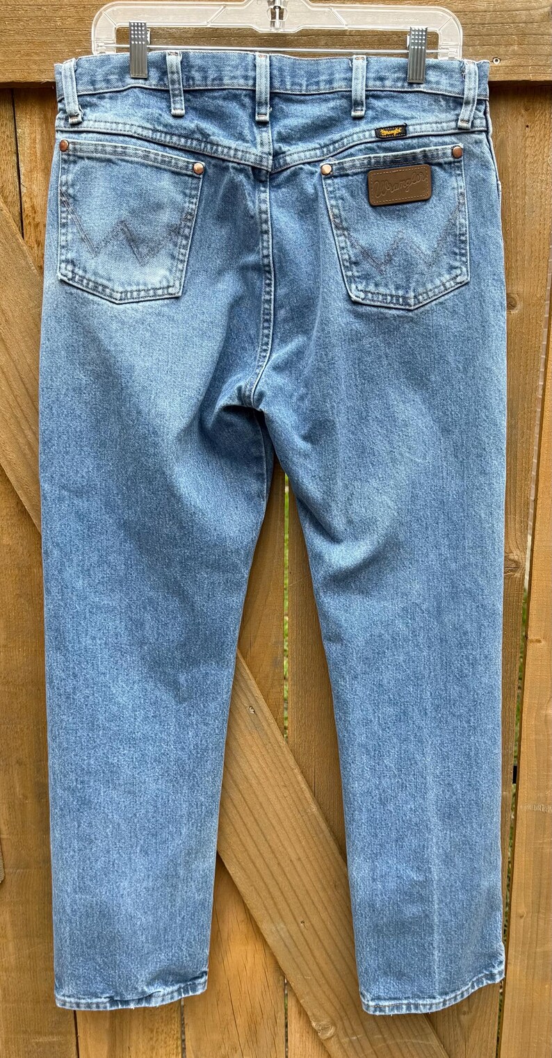 May include: Light blue denim jeans with classic five-pocket styling. The jeans feature a Wrangler brand patch on the back pocket and a metal button closure. The jeans are hanging on a hanger against a wooden background.