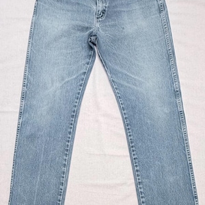 May include: Light blue denim jeans with a classic five-pocket design. The jeans have a straight leg cut and a button and zipper closure. The fabric appears to be pre-washed, giving them a faded look. The jeans are laid flat on a white surface.