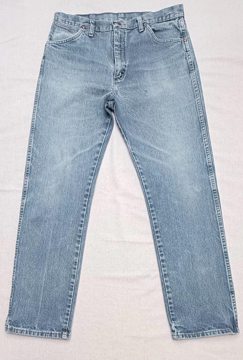 May include: Light blue denim jeans with a classic five-pocket design. The jeans have a straight leg cut and a button and zipper closure. The fabric appears to be pre-washed, giving them a faded look. The jeans are laid flat on a white surface.