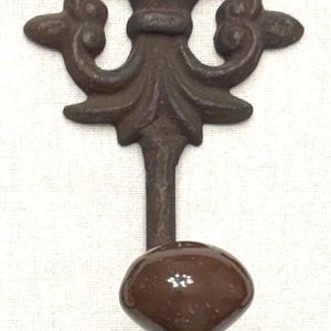May include: A wall-mounted hook featuring a dark brown, weathered metal backplate with an ornate, symmetrical scrollwork design and a circular loop at the top. A smooth, glossy, dark brown ceramic knob is attached to the front of the metal hook.