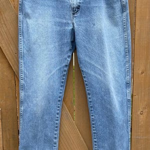 May include: Light blue denim jeans with a classic straight leg fit. The jeans are hung on a hanger against a wooden fence background. Features include a button closure, front pockets, and belt loops.