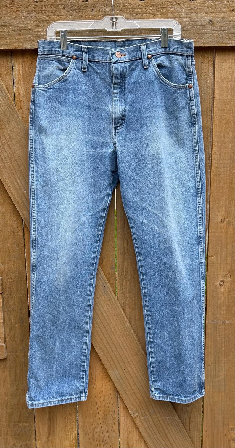 May include: Light blue denim jeans with a classic straight leg fit. The jeans are hung on a hanger against a wooden fence background. Features include a button closure, front pockets, and belt loops.
