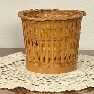 May include: A cylindrical woven basket made of light brown natural reeds. The basket features a vertical slat design with a reinforced rim and a solid base. The structure is open and airy, showcasing a consistent weave pattern throughout the body.