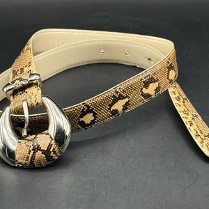 May include: A snakeskin-patterned belt with a silver-toned buckle. The belt features a beige interior and a brown and tan exterior with a repeating pattern. The buckle is a large, curved design with a snakeskin center.