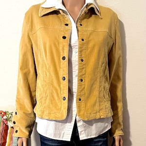 May include: Mustard yellow corduroy jacket with black buttons. The jacket has a collared neckline and long sleeves. A white button-down shirt is visible underneath. The jacket is paired with blue jeans.