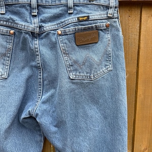 May include: Light blue denim jeans with copper rivets and a brown leather Wrangler patch on the back pocket. The jeans are hanging on a metal hanger. The Wrangler logo is visible on the patch and above the back pocket.